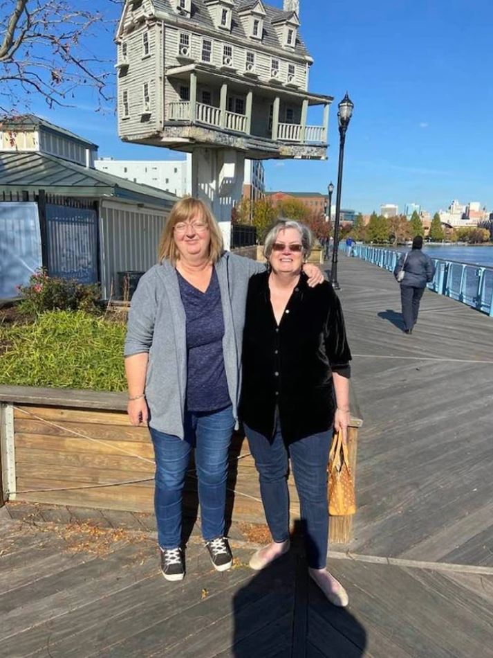 Dve ženy na promenáde s surrealistickým domom na stĺpe v pozadí za slnečného dňa.