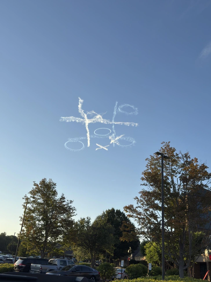 Krížiky a krúžky na oblohe vytvorené dymovými čiarami nad parkoviskom a stromami.