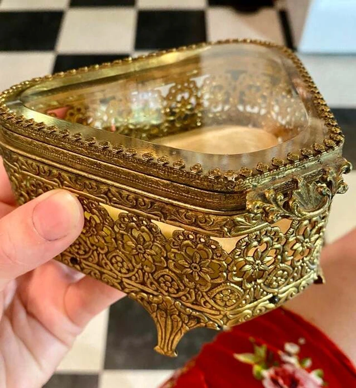 Zdobená mosadzná truhlička s priehľadným vrchnákom, držiac ju na ruke na šachovnicovej podlahe.