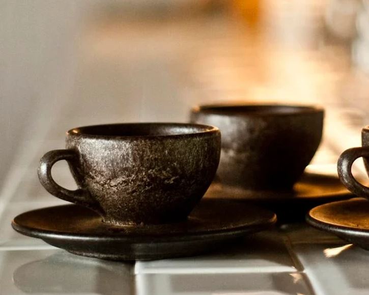 Tmavé keramické šálky na kávu na tanierikoch, osvetlené teplým svetlom.