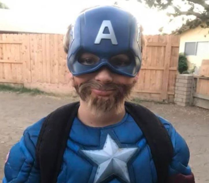 Dieťa v kostýme superhrdinu s modrou maskou a falošnou bradou stojí pred dreveným plotom.