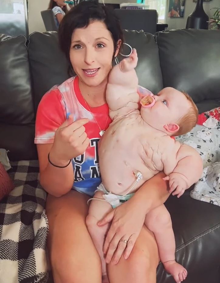 Loísa s bábätkom na pohovke, s úsmevom, drží dieťa v náručí, ktoré má cumlík. Domáce prostredie, gauč.