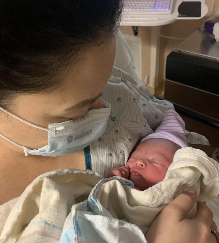 Matka v nemocničnej izbe drží novorodenca v náručí, obaja sú v ochranných rúškach.