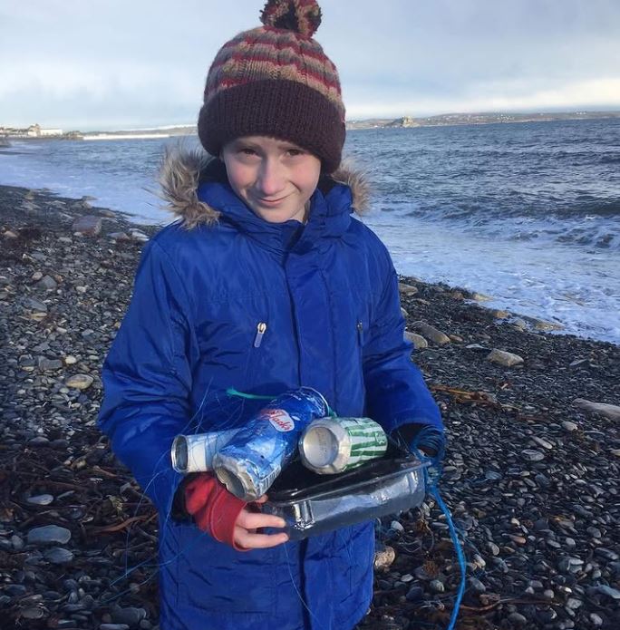 Mladý chlapec v modrej bunde zbiera odpadky na kamenistej pláži pri mori.