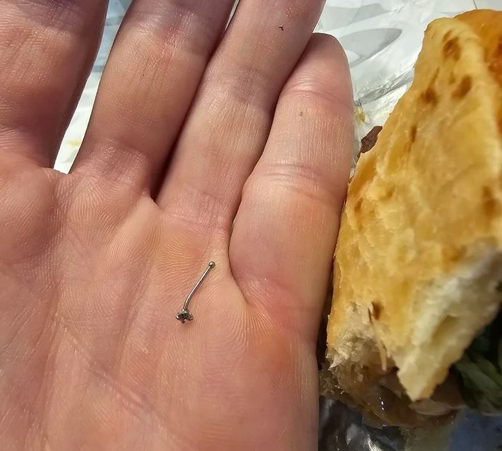 Ruka držiaca malý kovový predmet, vedľa je sendvič na stole.