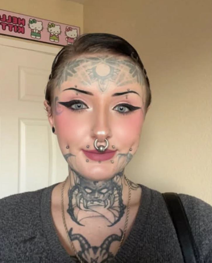 Osoba s výraznými tetovaniami a piercingami na tvári pózuje v interiéri, v pozadí dekór s motívom Hello Kitty.