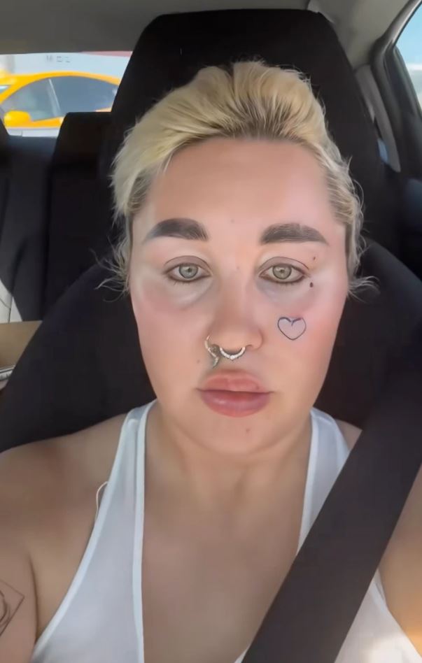 Osoba s blond vlasmi a tetovaním srdca na tvári sedí v aute, na sebe má biele tielko.