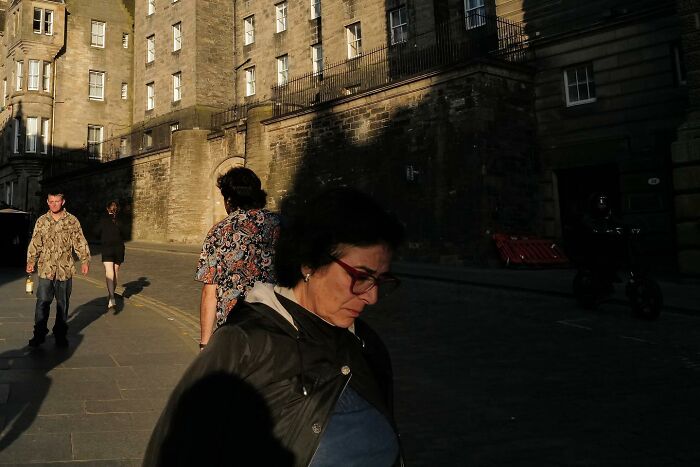 Žena prechádza tienistou ulicou s historickou budovou v pozadí, usilovne kráča v podvečernom slnku.