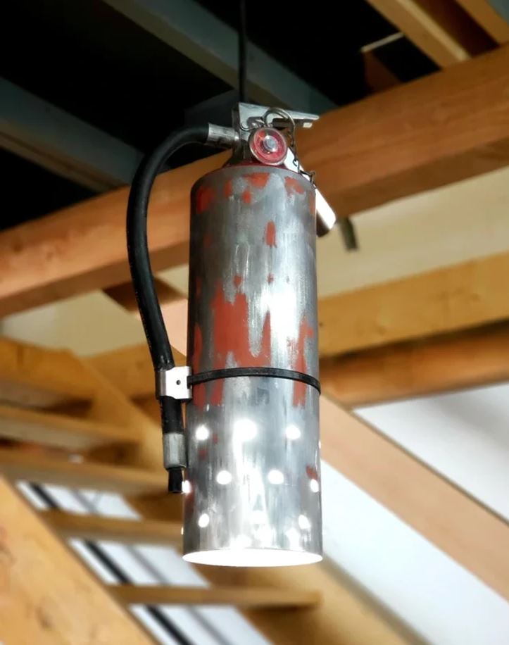 Priemyselná loftová lampa z recyklovaného hasičského prístroja visí na strope z drevených trámov.