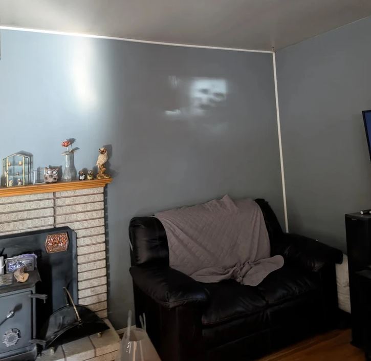 Obývačka s čiernym koženým kreslom, šedou dekou a svetlom na stene, vedľa kachlí a dekorácií na krbe.