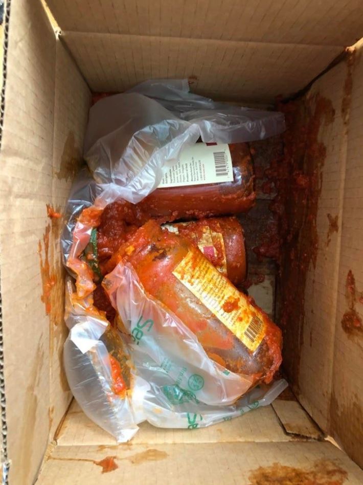 Rozbitá sklenená fľaša s paradajkovou omáčkou v kartónovej krabici, rozliaty obsah.