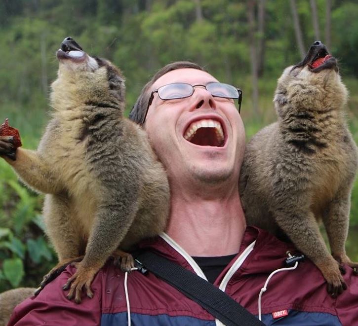 Muž so širokým úsmevom, s dvoma lemurmi na ramenách v prírodnom prostredí.