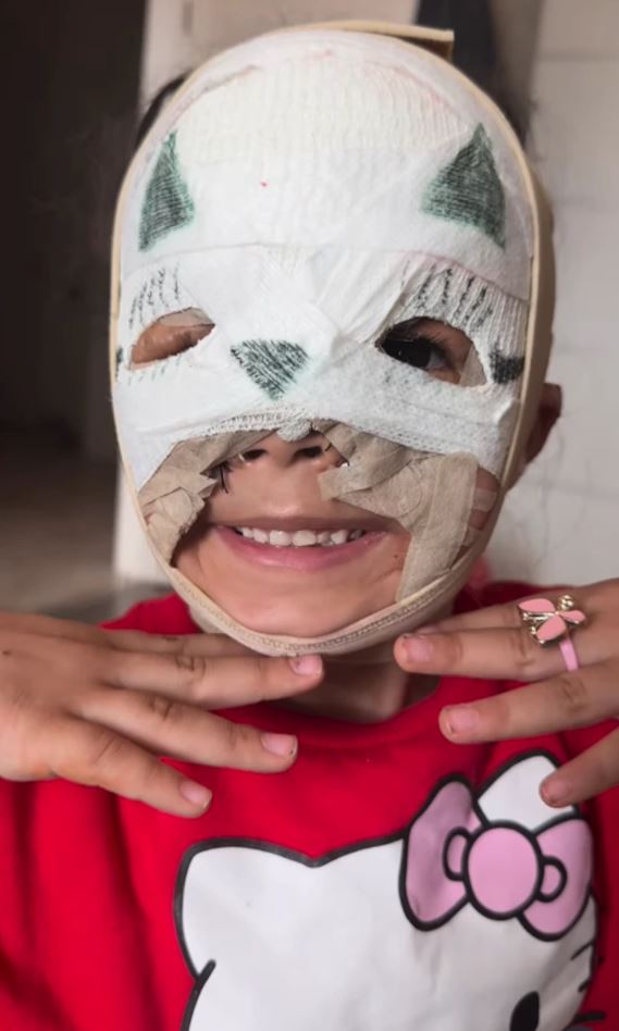 Dieťa s úsmevom nosí ručne vyrobenú masku na tvár, červené tričko s mačacím motívom.