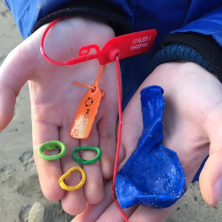 Ruky držiace farebné plastové predmety nájdené na pláži: krúžky, úlomky a modrý balónik.