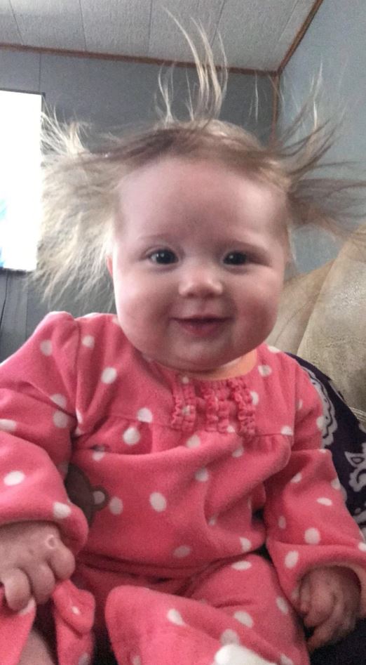 Roztomilé dieťa v ružovom oblečení s rozstrapatenými vlasmi sedí na pohovke a usmieva sa do fotoaparátu.