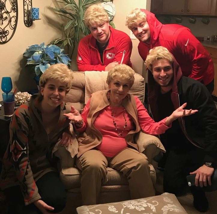 Skupina ľudí v kostýmoch s parochňami, usmiati v obývačke okolo sediacej ženy na kresle.