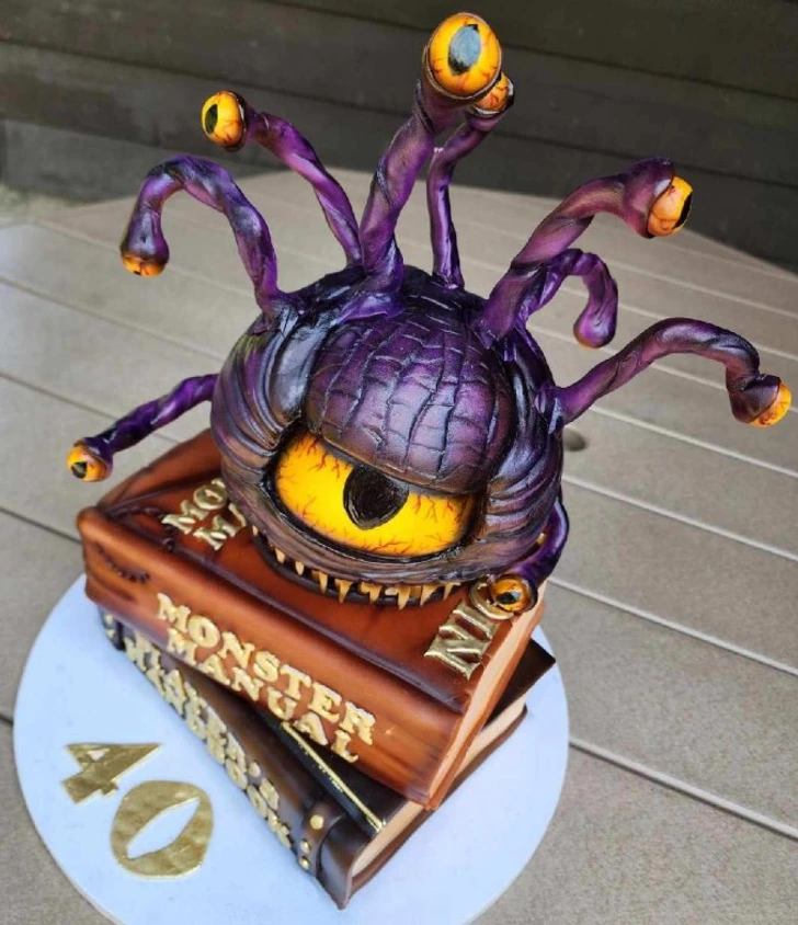 Fialovo-žltá monštrum torta s očami na vrchu knihy Monster Manual, oslava 40. narodenín.