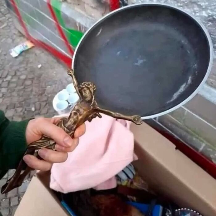 Osoba drží neobvyklý predmet: panvicu s rukoväťou v tvare bronzového krucifixu na trhovisku.