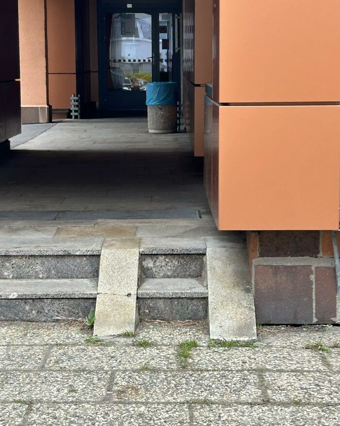 Rampy a schody vedúce k dverám moderného oranžového budovy s modrým košom na odpadky.