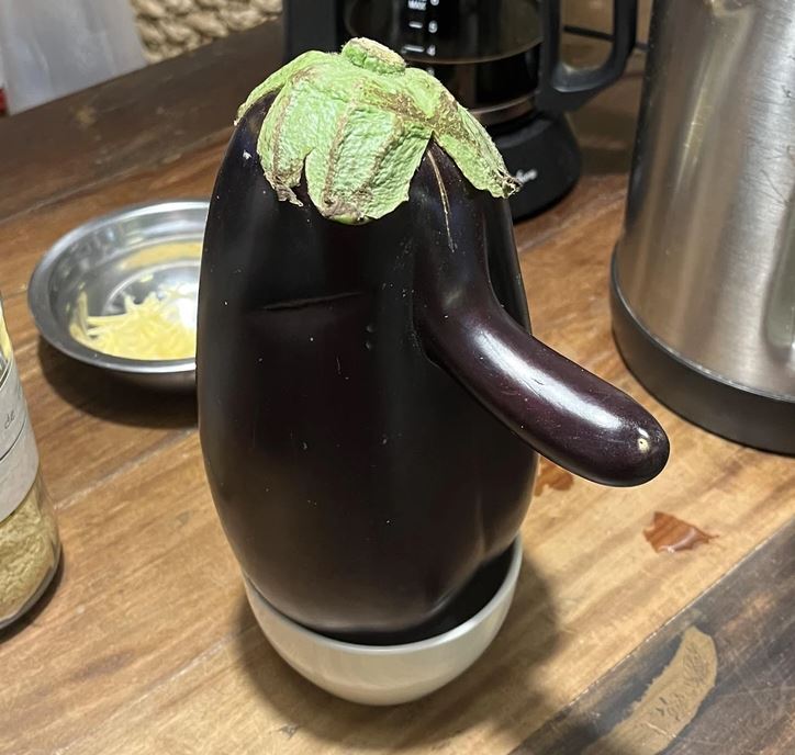 Fialový baklažán položený na stole s tvarom pripomínajúcim tučniaka, detail kuchynskej linky v pozadí.