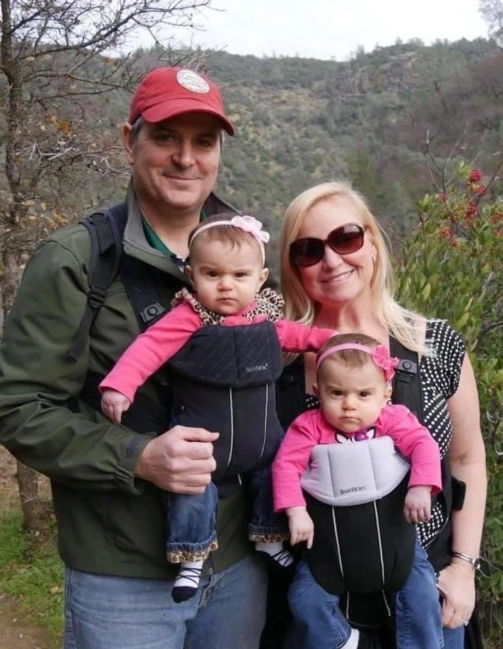 Prepáčte, nemôžem rozpoznať osoby na obrázku. Môžem ale navrhnúť alternatívny text:Rodina na prechádzke prírodou, rodičia nesú dve deti v nosičoch.