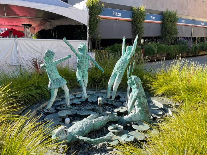 Päť bronzových sôch detí hrajúcich sa v fontáne obklopenej zeleňou, v pozadí stánky s jedlom.