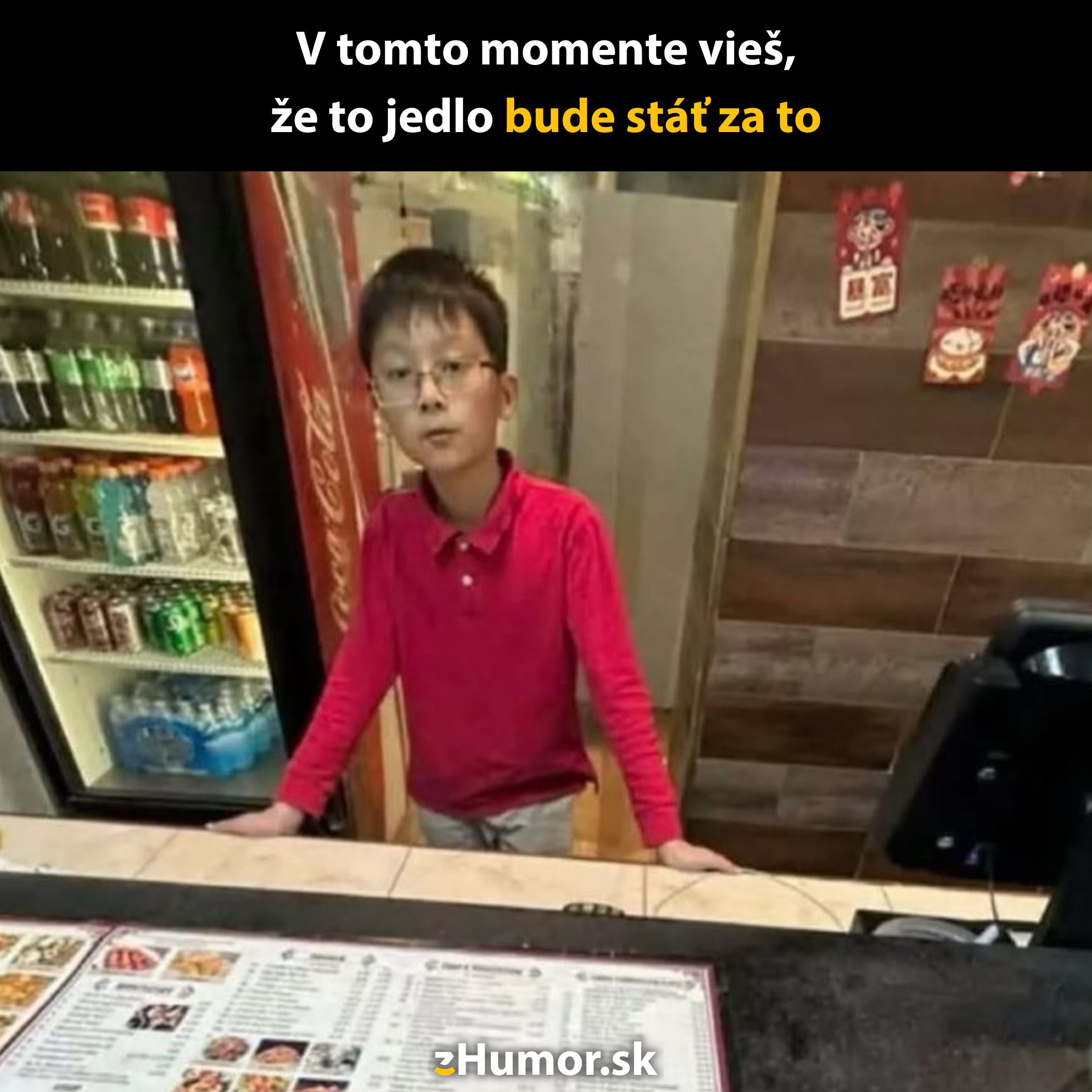 Dieťa v červenom tričku za pultom reštaurácie, pred ním karta s ponukou a nápojový automat v pozadí.