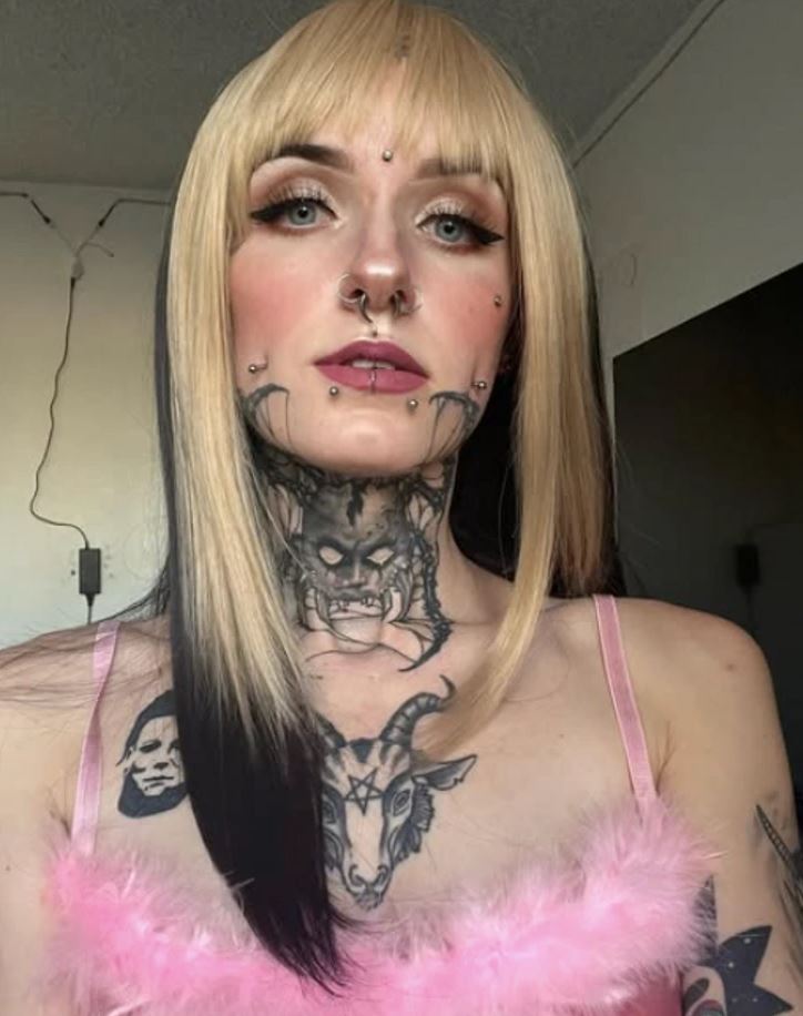 Mladá žena s blond vlasmi, výrazným make-upom a tetovaniami na krku a tvári, pózuje v interiéri.