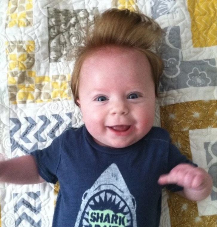 Usmievavé bábätko s vysokým účesom v modrom tričku s potlačou žraloka na vzorovanej prikrývke.