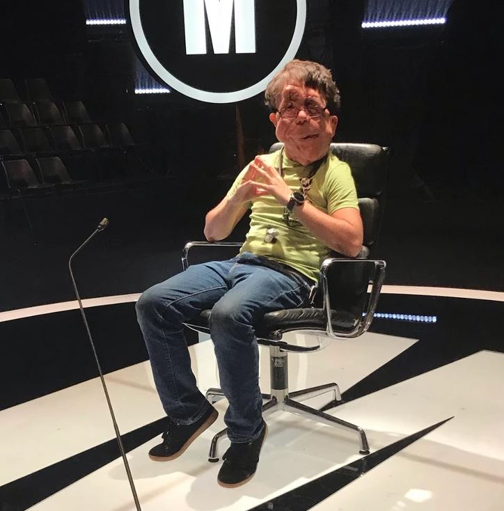 Osoba sedí na scéne v kresle s mikrofónom, oblečená v zelenom tričku a džínsach.