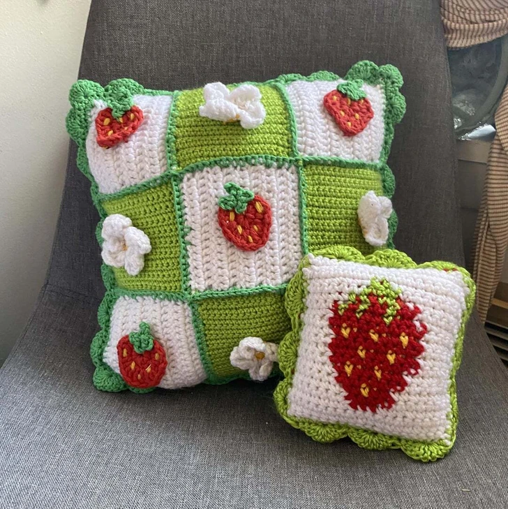 Háčkované vankúše s jahodovým vzorom na šedom kresle, dekorované kvetmi a zelenými okrajmi.