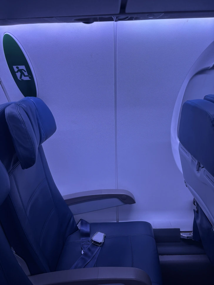 Sedadlo v lietadle vedľa núdzového východu osvetlené modrým svetlom, pás pripravený na použitie.