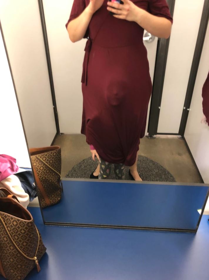 Osoba skúša bordové šaty v kabínke obchodu pred zrkadlom, vedľa je hnedá kabelka.