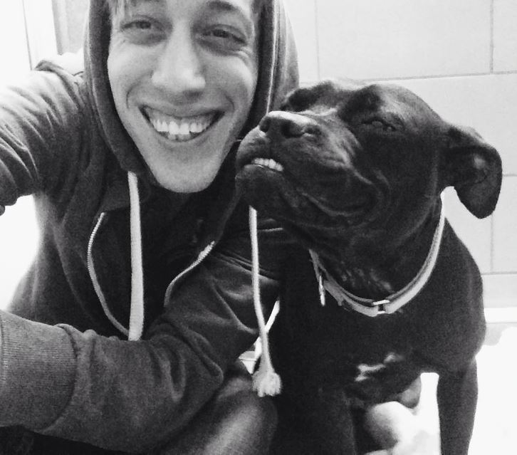 Osoba v mikine sa usmieva spolu s čiernym psom, obaja vyzerajú šťastne.