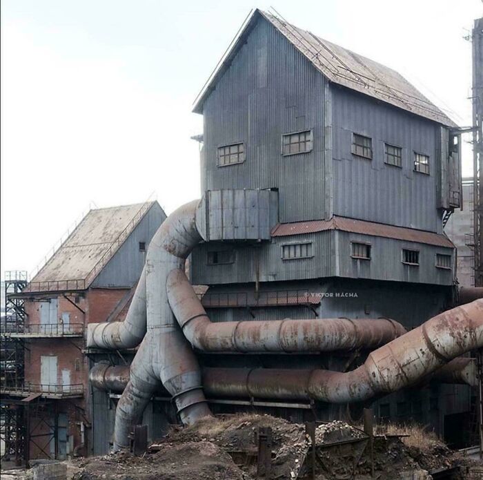 Stará priemyselná budova s kovovými potrubiami a plechovou strechou v opustenom výrobnom areáli.