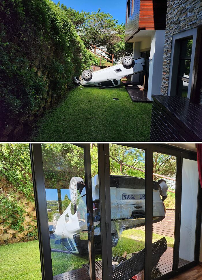 Prevrátené auto na záhrade naráža do domu, rozbitá sklenená stena, dramatická nehoda v pokojnom prostredí.