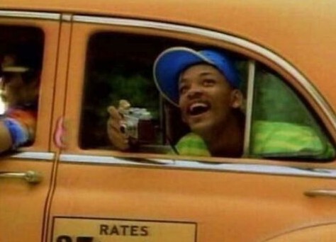 Osoba v aute s fotoaparátom vyzerá nadšene a pohodlne, pozerá z okna staršieho vozidla oranžovej farby.