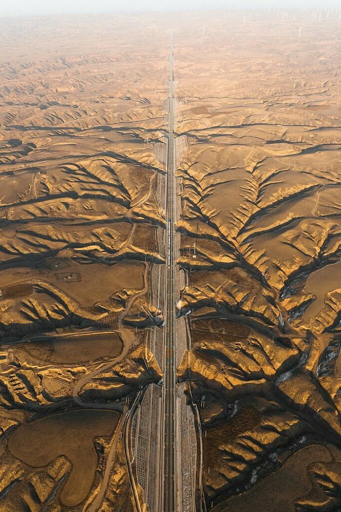 Letecký pohľad na diaľnicu prechádzajúcu púštnou krajinou s veternými turbínami na horizonte.
