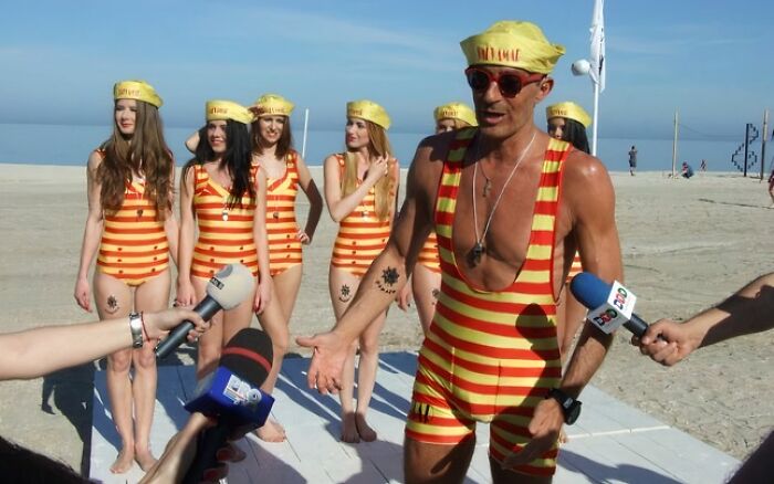 Muž v pruhovanom oblečení a klobúku hovorí s novinármi na pláži, lemovaný ženami v podobných plavkách.