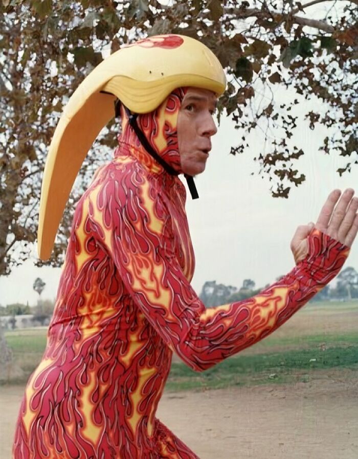 Muž v plamenistom kostýme s helmou v tvare vtáka beží vonku pri stromoch.