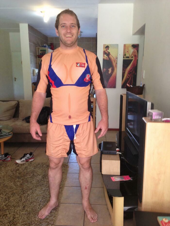 Muž s úsmevom v zábavnom kostýme, ktorý vyzerá ako telo s bikinkou, stojí v obývačke.