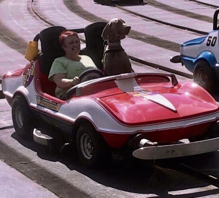 Žena s hnedým psom na okružnej dráhe v červenom autíčku, obaja nadšene sledujú jazdu.