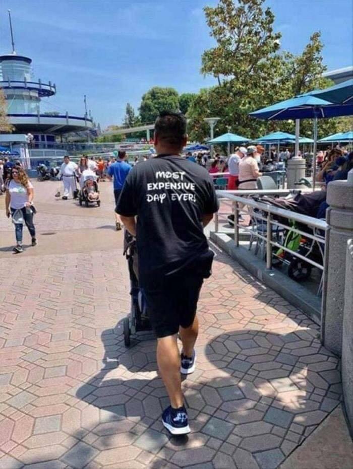 Muž v tričku Najdrahší deň vôbec tlačí kočík v zábavnom parku plnom ľudí a slnečníkov.