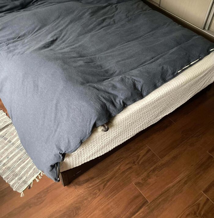 Tmavá posteľná prikrývka na posteli na drevenej podlahe; textúrovaný koberec vedľa postele.