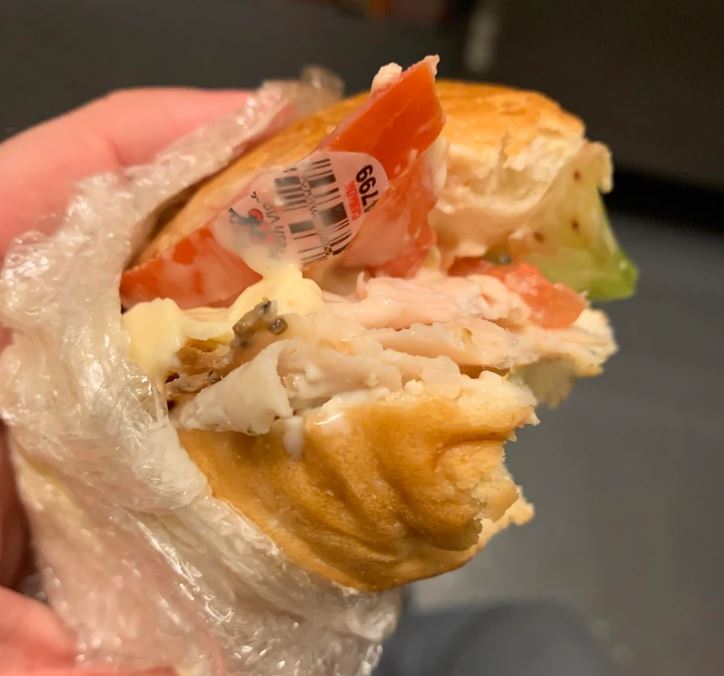 Obrázok rozbaleného sendviča s nálepkou na rajčine, držaného v ruke, pripravený na konzumáciu.