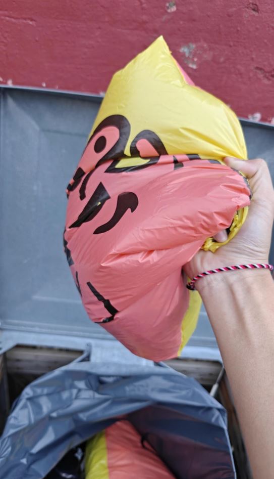 Osoba drží plastové vrece s odpadkami pred kontajnerom na odpadky.