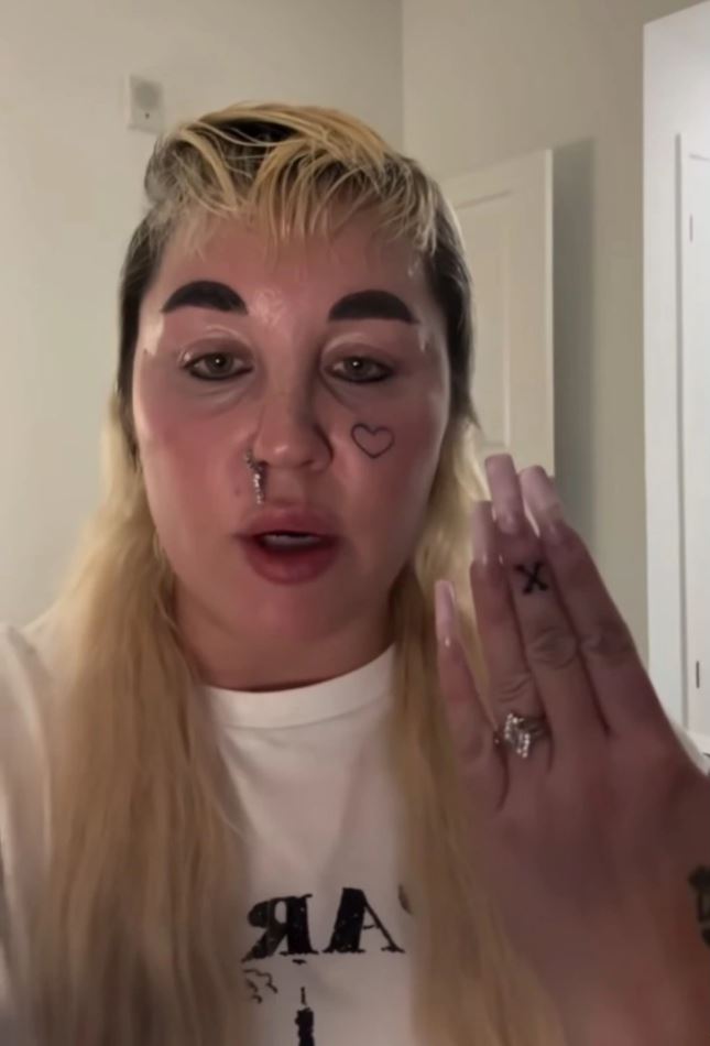 Osoba s tetovaním srdca na tvári ukazuje rukou s prsteňom. Má blond vlasy a biele tričko.