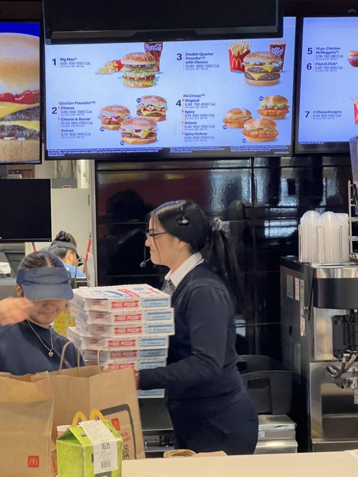 Zamestnankyňa fastfoodu nesie krabice pri pokladni, na pozadí ponuka jedál na obrazovkách.