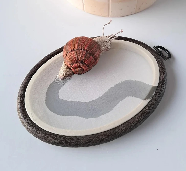 Vyšívaný slimák na oválnej drevenej výšivke, vytvára ilúziu pohybu na textilnom plátne.