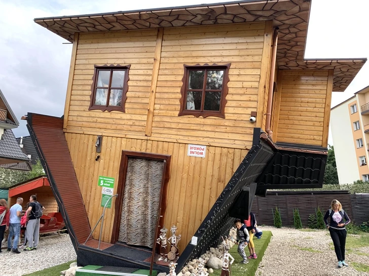 Obrátený dom so strechou na zemi a podlahou hore, návštevníci si ho zvedavo prezerajú.